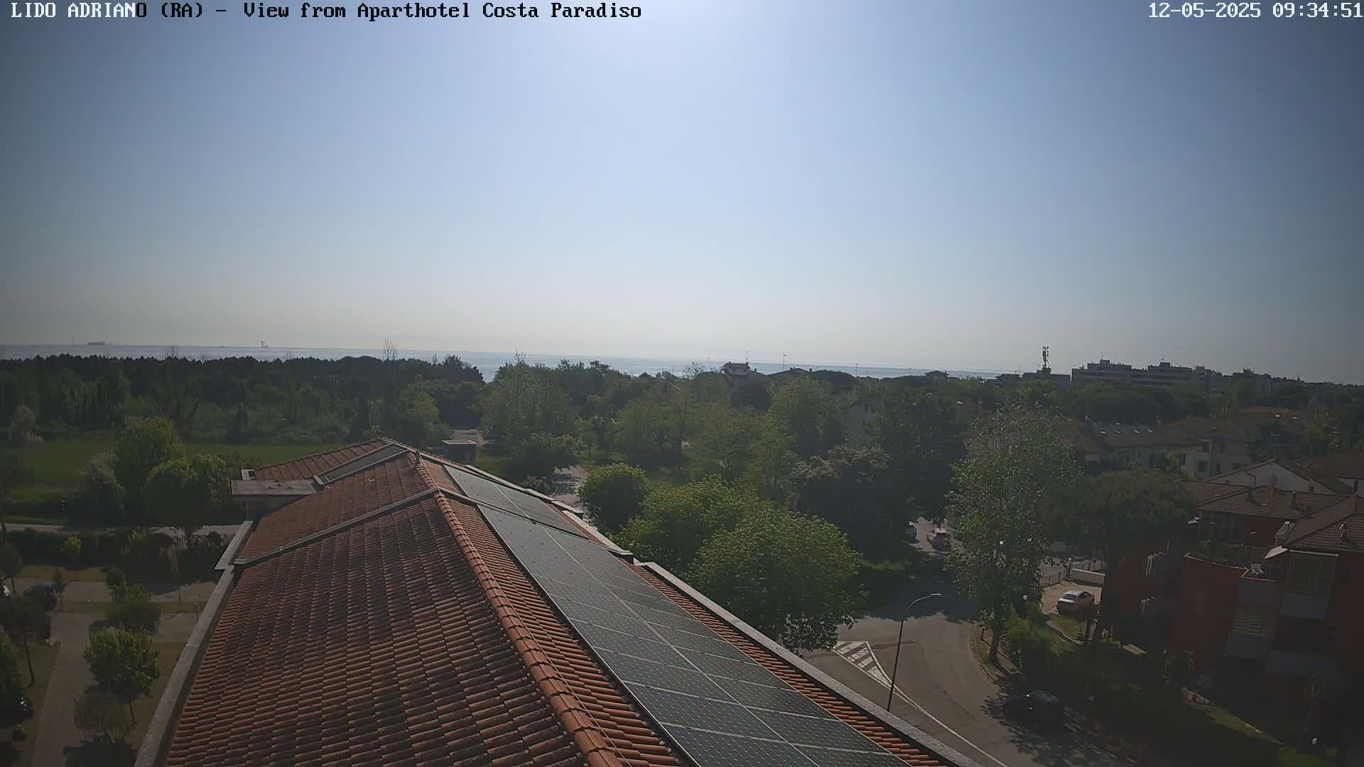 A rooftop with solar panels overlooks a clear, sunny day with a calm sea and green tree-lined streets in the distance.