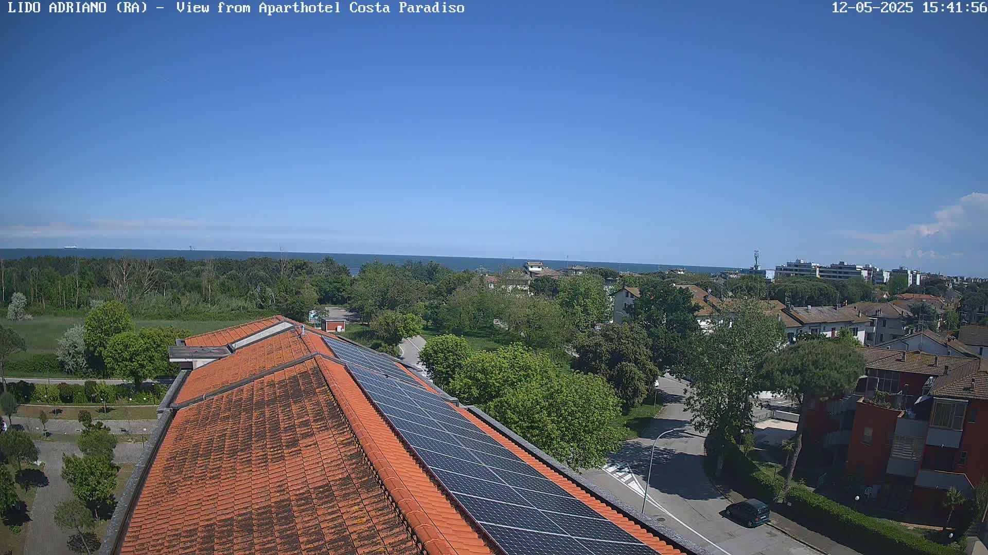 A clear, sunny day overlooks an orange-tiled roof with solar panels, a residential area with green trees, and a calm ocean in the distance.