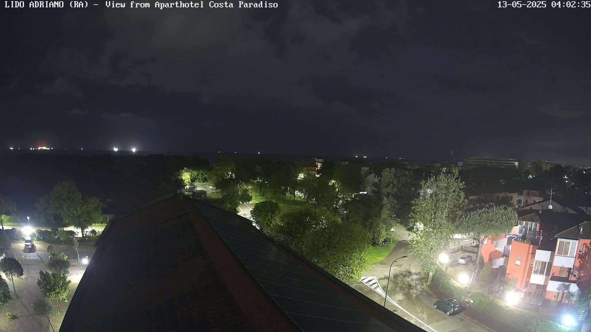 A nighttime, overcast view of a residential area with trees and buildings illuminated by streetlights, showing wet roads and a partially visible rooftop in the foreground.