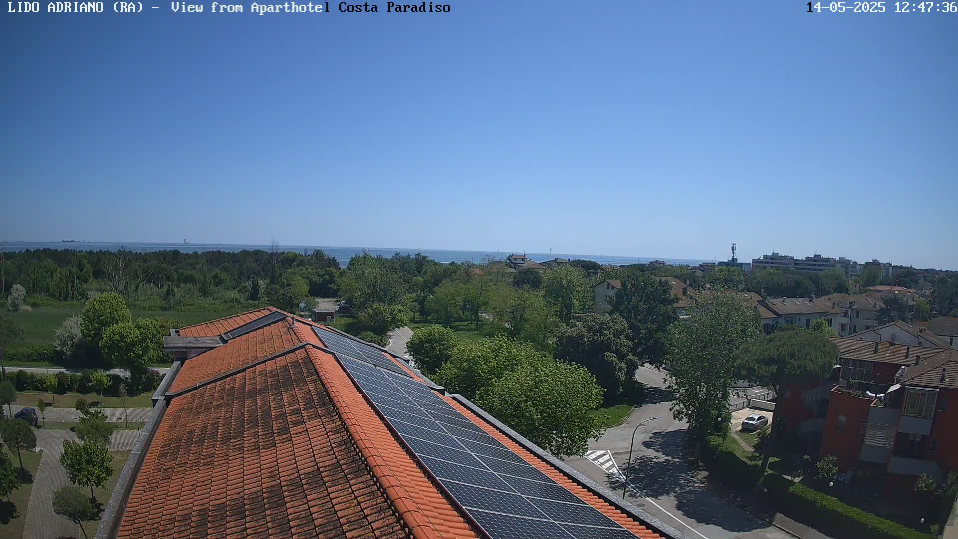 A clear, sunny day overlooks a coastal town with a red-tiled roof and solar panels visible in the foreground, showcasing a tranquil seaside scene.