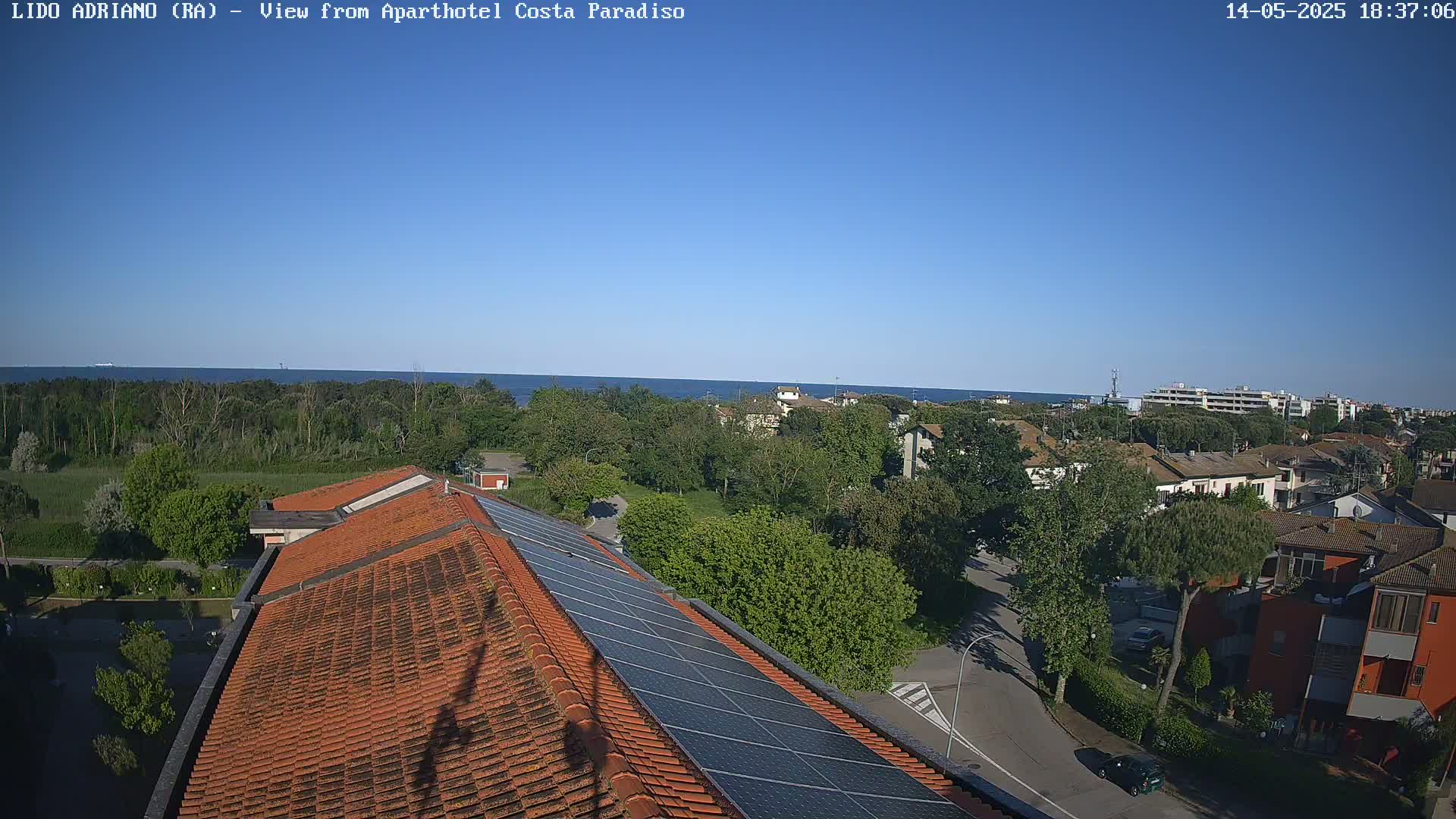 A clear, sunny day overlooks a coastal town with red-tiled roofs and solar panels, a calm sea visible in the distance.