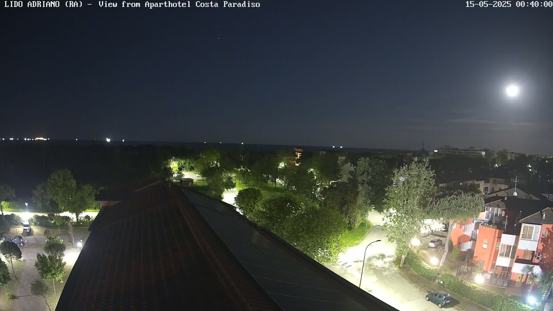 A nighttime view from a rooftop shows a moonlit seaside town with a calm body of water in the distance, illuminated streets, and buildings surrounded by green trees.
