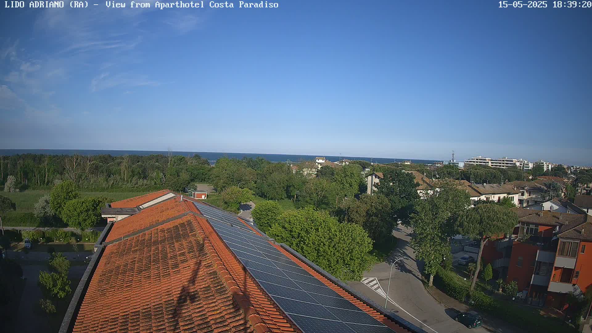 A clear, sunny day overlooking a coastal town with a tiled roof and solar panels in the foreground, and a calm ocean in the distance.