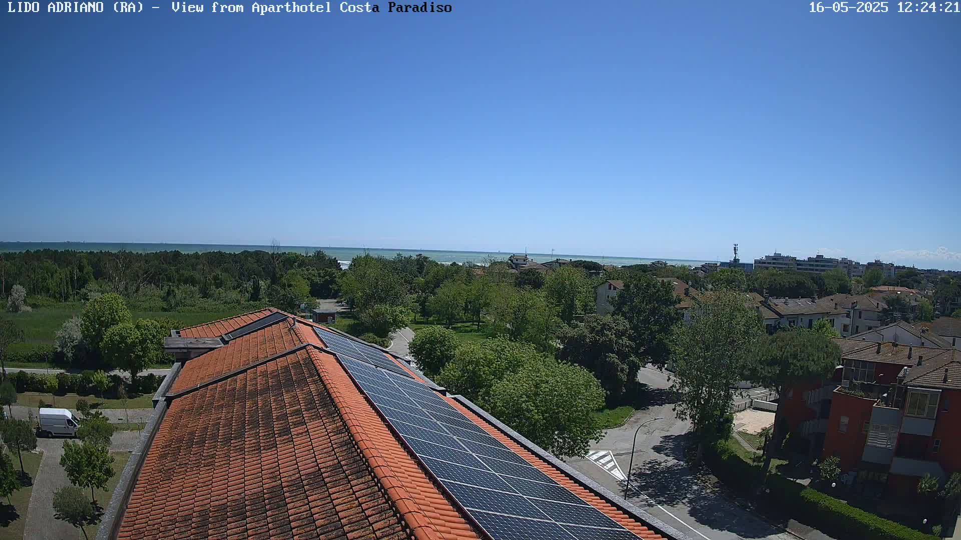 A rooftop with solar panels overlooks a residential area, lush green trees, and a calm ocean under a clear blue sky.