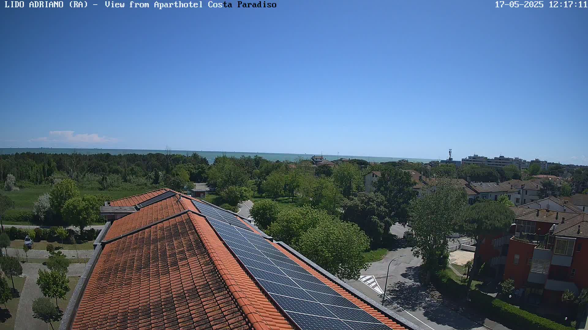 A rooftop with solar panels overlooks a residential area and a calm blue sea under a clear sky.