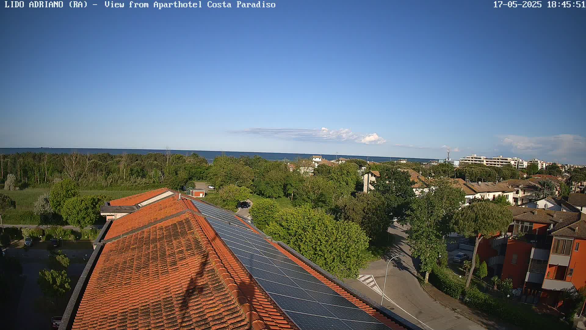 A mostly clear blue sky overlooks a coastal town with red-tiled roofs and solar panels on a building, trees and a calm ocean in the distance.