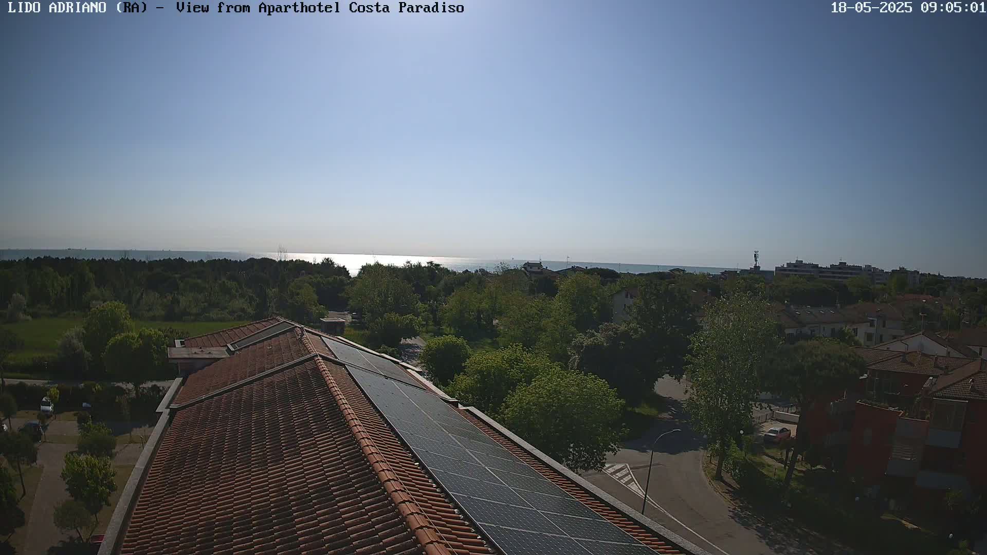 A rooftop view of a building with solar panels, overlooking a calm ocean and a tree-lined residential area under a clear, sunny sky.