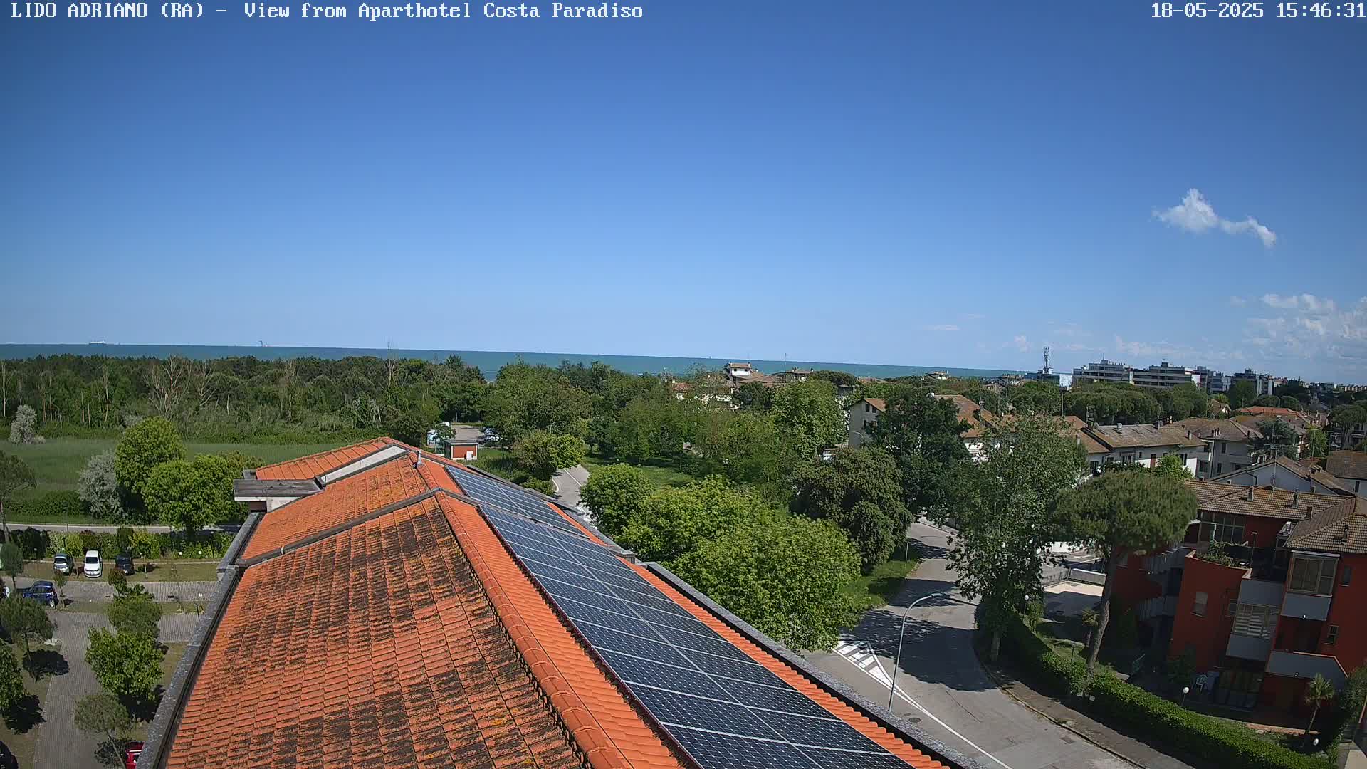 A mostly sunny day reveals a view of a seaside town from a rooftop with solar panels, showing a calm sea in the distance and lush green trees and buildings in the foreground.