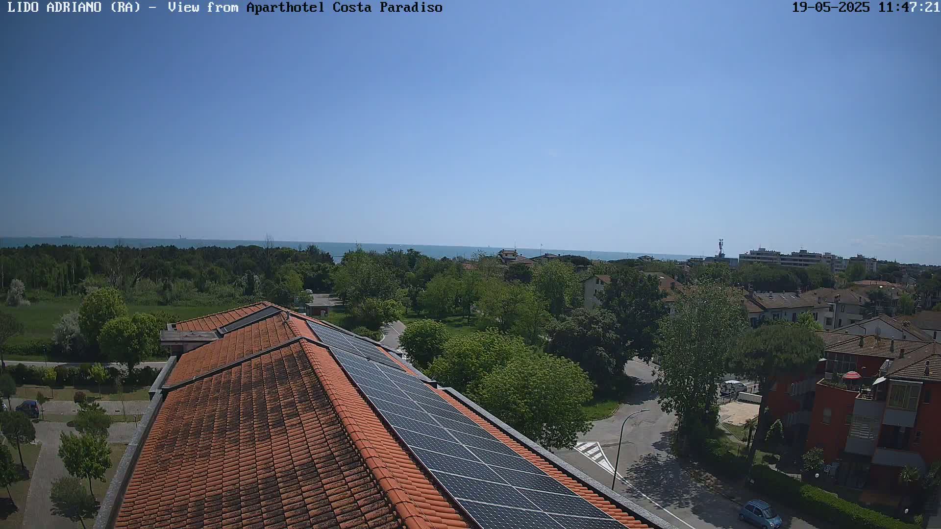 A building with a red tile roof and solar panels is situated overlooking a residential area with green trees and a calm blue sea in the distance under a clear, sunny sky.
