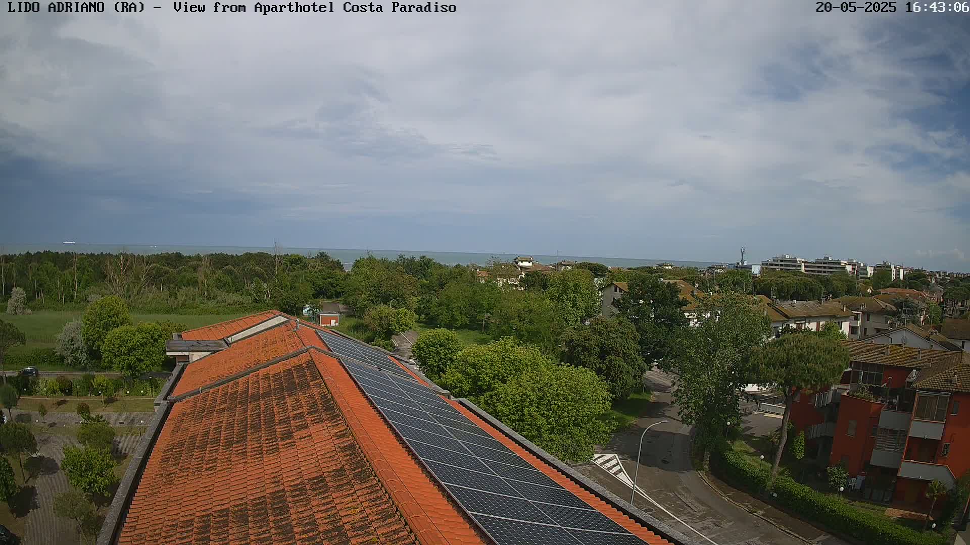 An aerial view shows a partly cloudy sky over a coastal town with red-tiled rooftops, lush green trees, and a calm ocean in the distance.