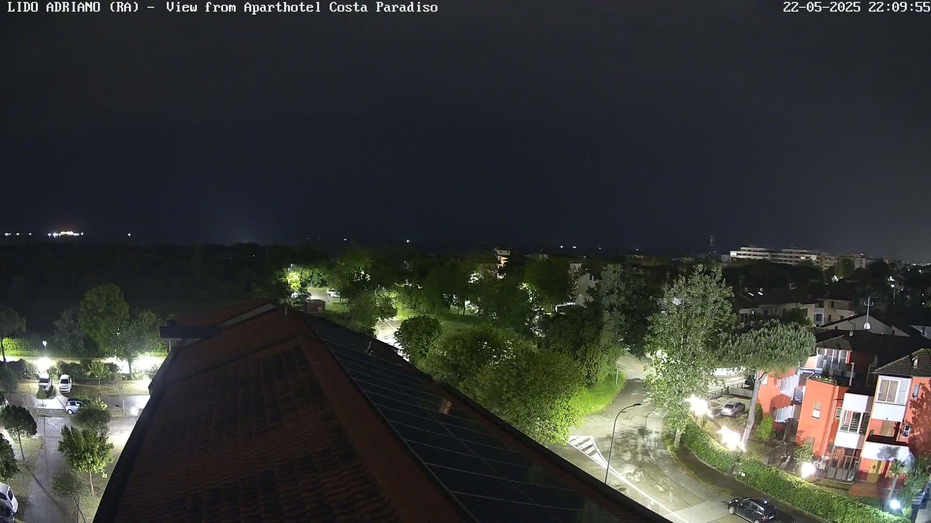 A nighttime, rain-slicked view of a residential area with a partial view of a building's roof with solar panels, surrounded by trees and streetlights.