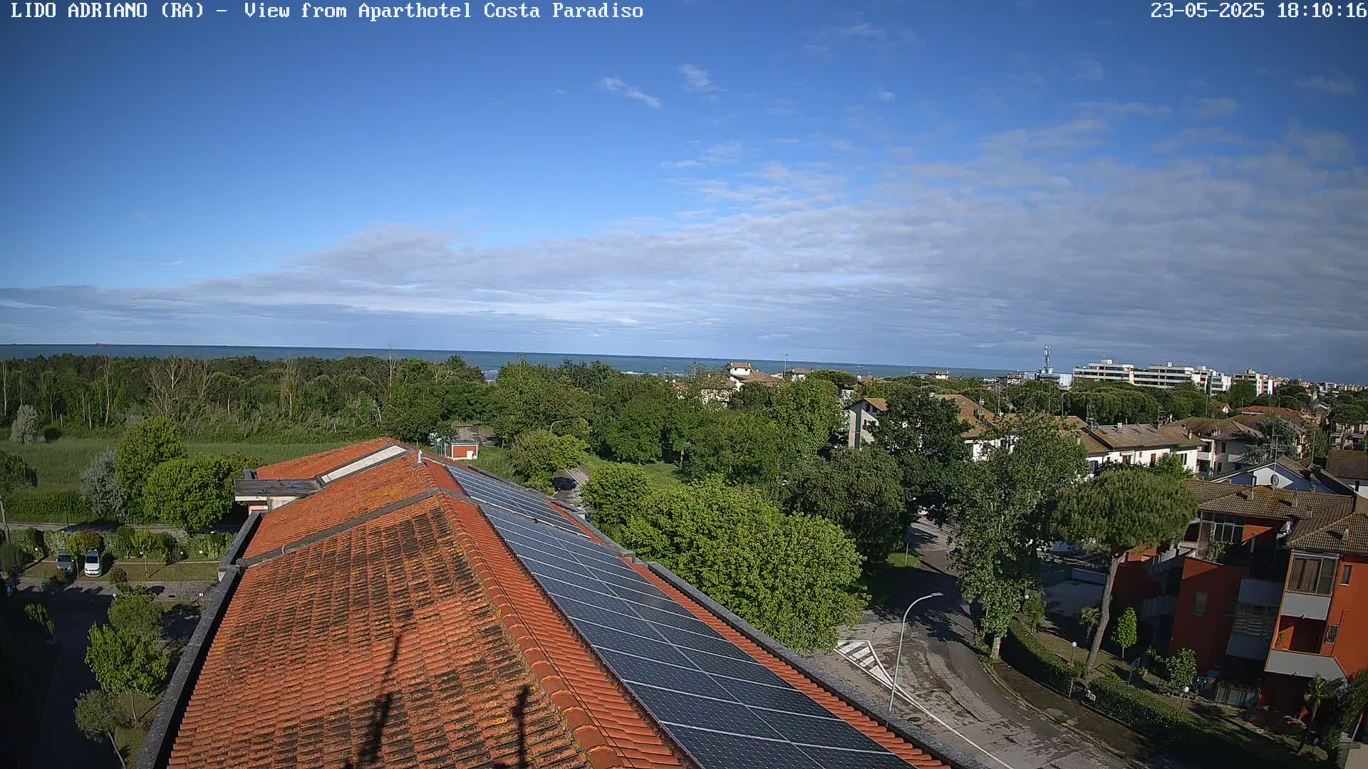 A partly cloudy sky overlooks a coastal town with a red tiled roof building featuring solar panels, green trees, and a calm ocean in the distance.