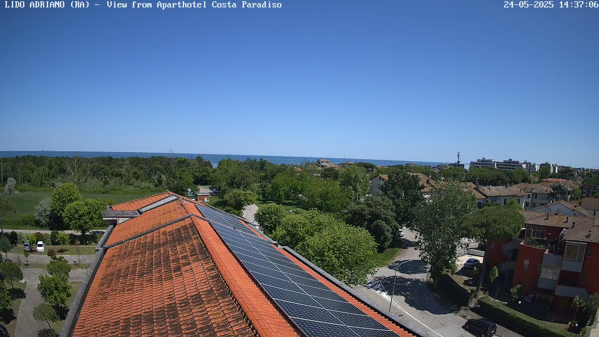 A building with an orange tile roof and solar panels overlooks a tree-lined residential area and a calm blue sea under a clear sky.
