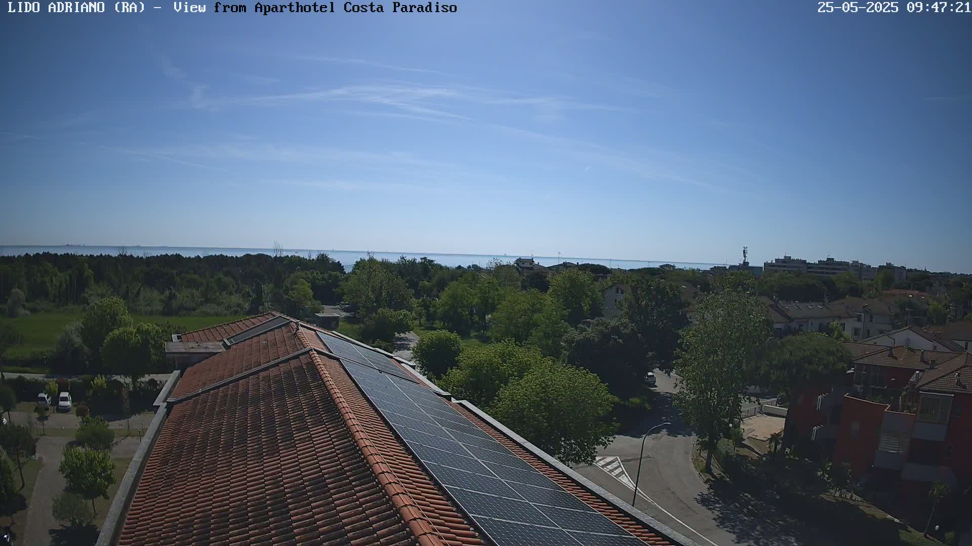 A rooftop with solar panels overlooks a residential area and a calm sea under a clear blue sky.