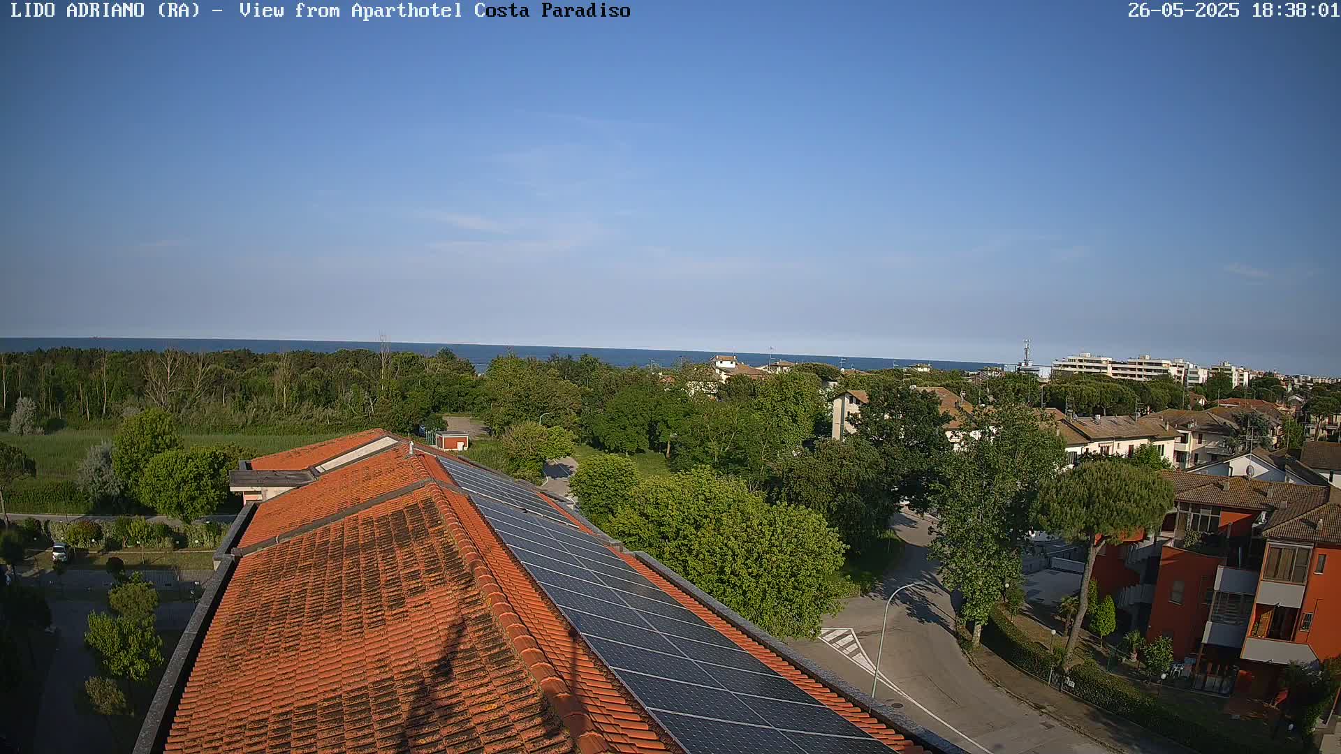 A clear, sunny day overlooks a coastal town with a reddish-brown tiled roof building featuring solar panels, and a calm ocean in the distance.