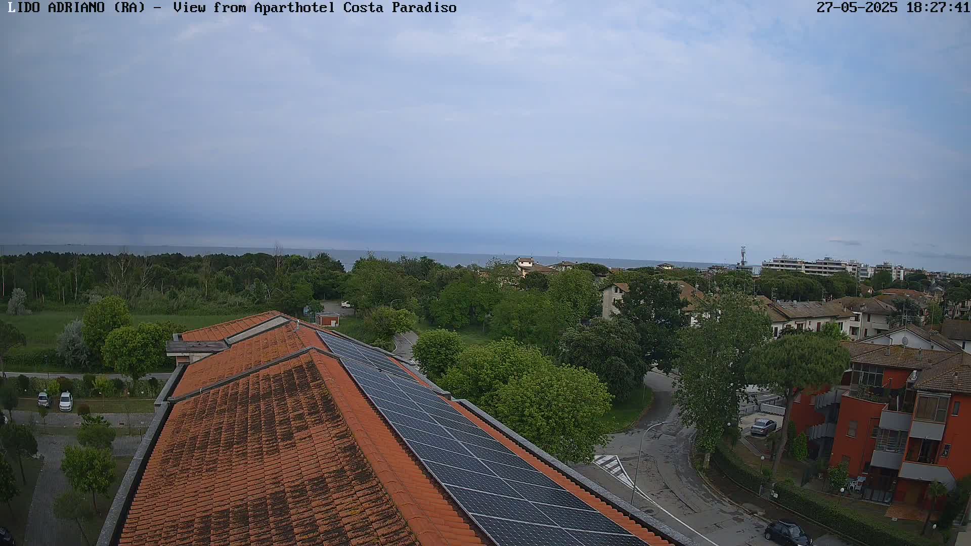 An aerial view shows a coastal town under a mostly cloudy sky, with a tiled roof and solar panels in the foreground and the sea visible in the distance.