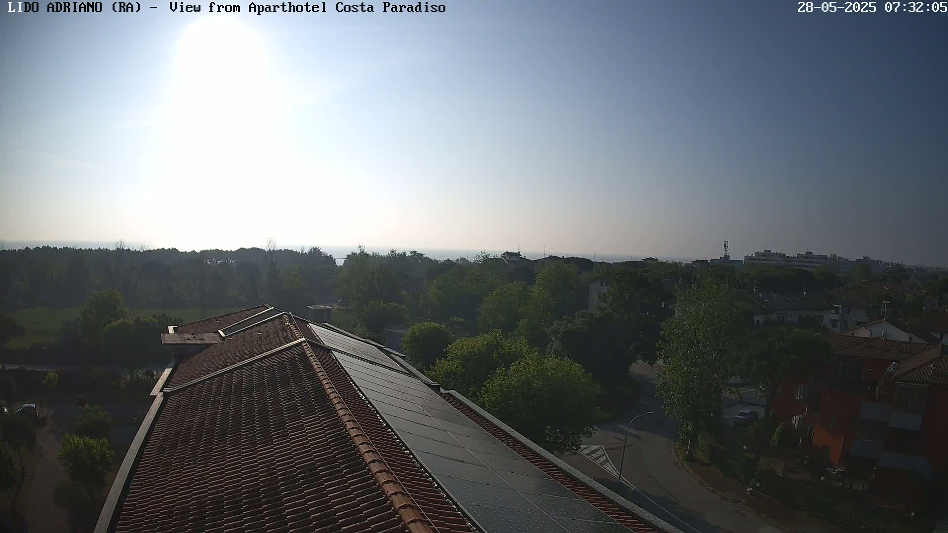 A high-angle view shows a red-tiled roof with solar panels, overlooking a tree-lined suburban area and a calm body of water under a clear, bright sky.