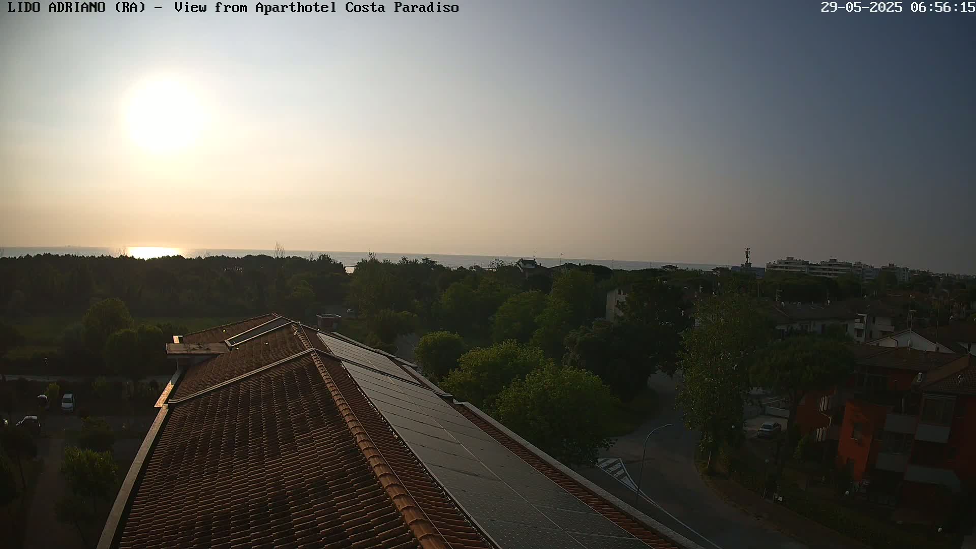 A sunlit coastal scene is viewed from a rooftop with solar panels, showing a calm sea, green trees, and residential buildings in the late afternoon or early evening.