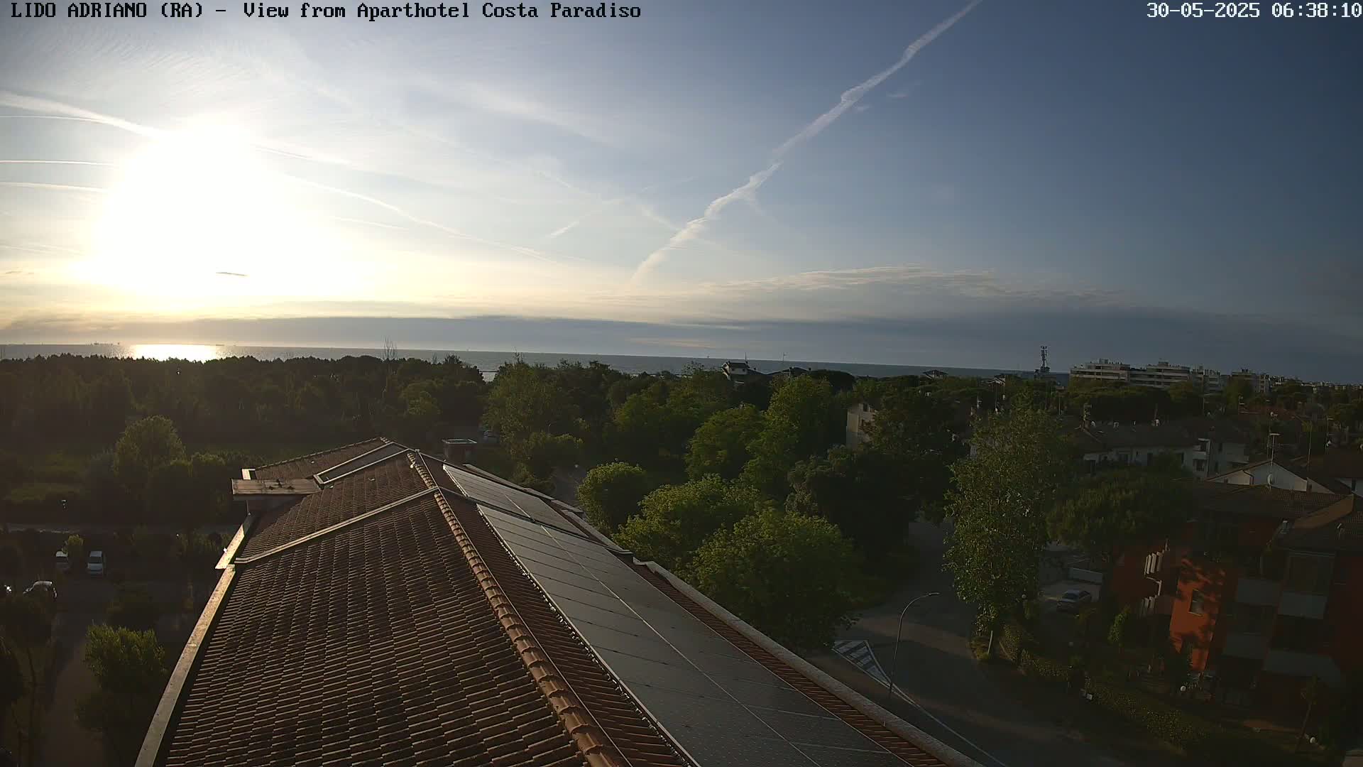 A sunrise over a calm sea is visible from a rooftop with solar panels, overlooking a tree-lined residential area.