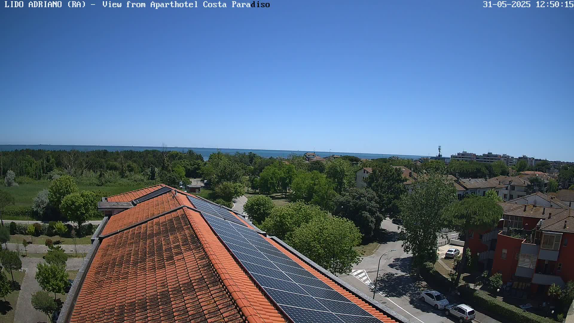 A clear blue sky overlooks a red-tiled rooftop with solar panels, a residential area with green trees, and a calm ocean in the distance.