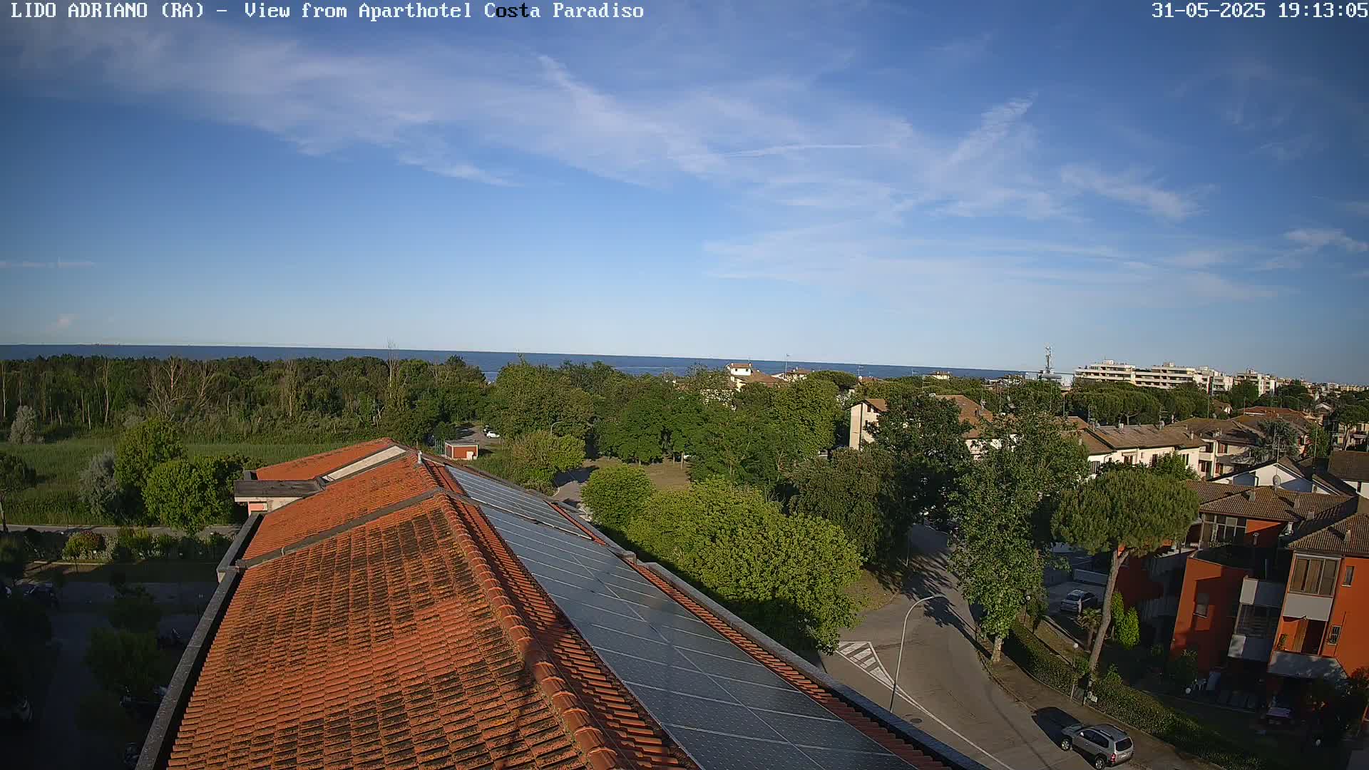 An aerial view shows a mostly sunny day overlooking a coastal town with a calm sea in the distance, featuring red-tiled rooftops and solar panels.