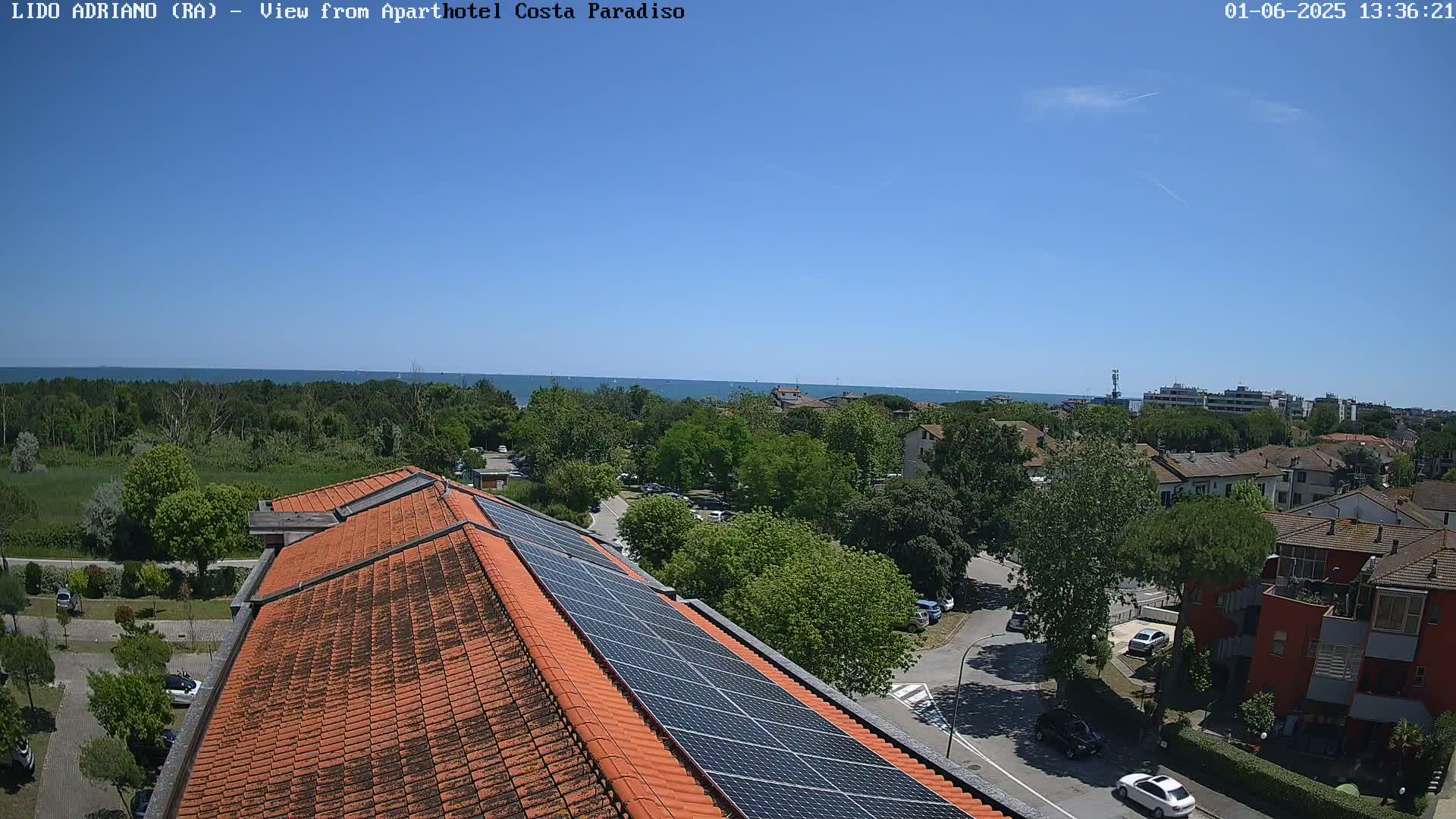 A rooftop with solar panels overlooks a residential area with lush green trees, a calm blue sea in the distance, under a clear, sunny sky.