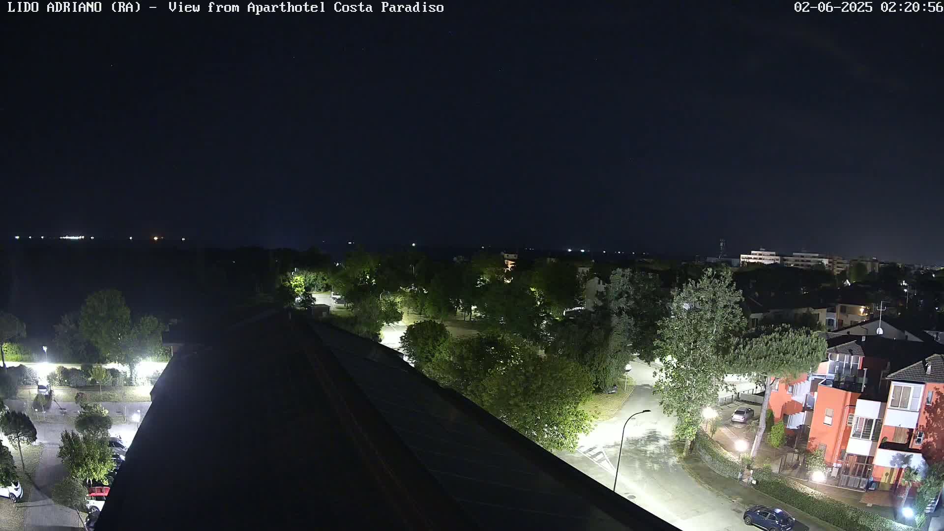 A nighttime, overhead view of a residential area with streetlights illuminating roads, trees, and buildings near a body of water under a clear, dark sky.