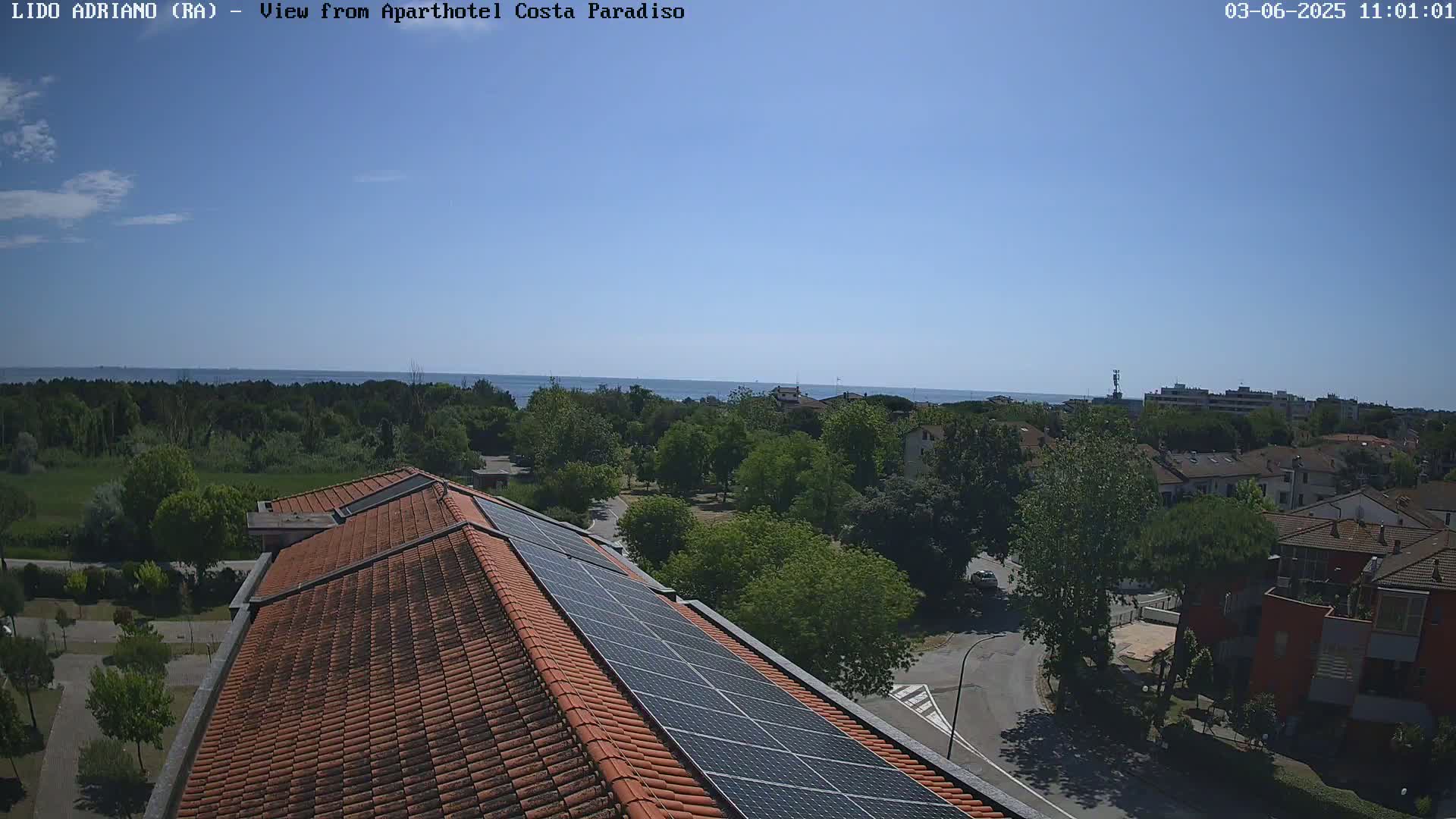 A rooftop with solar panels overlooks a tree-lined residential area and a calm sea under a clear blue sky.