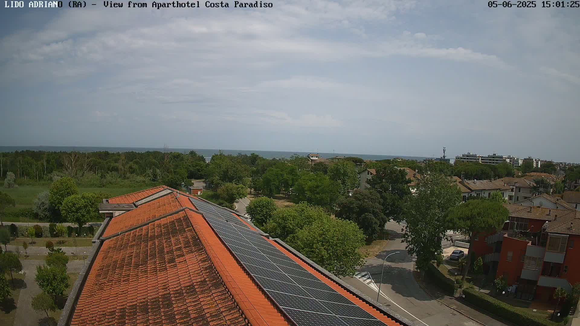 A partly cloudy day overlooks a coastal town with a red-tiled roof building featuring solar panels, and a calm sea visible in the distance.