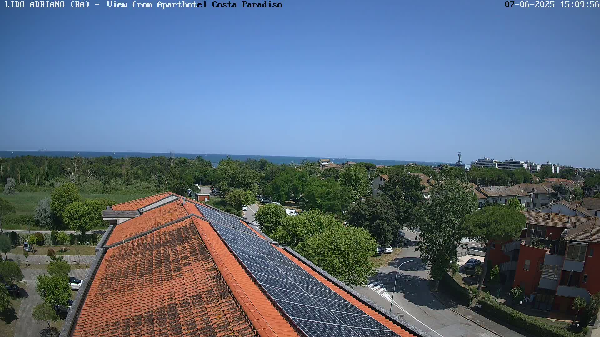 A rooftop with solar panels overlooks a residential area and a calm ocean under a clear blue sky.