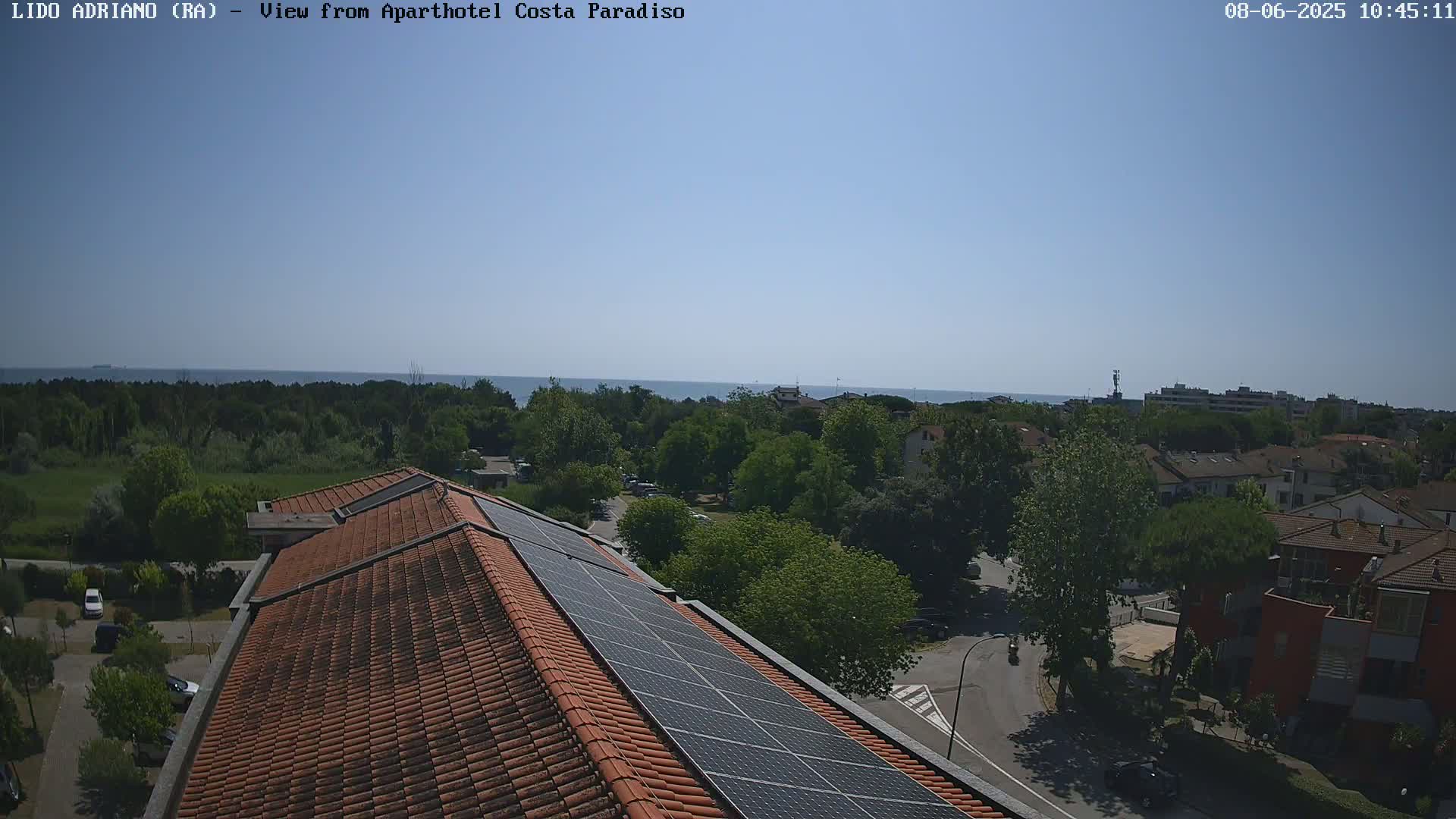 A rooftop with solar panels overlooks a residential area and a calm sea under a clear blue sky.