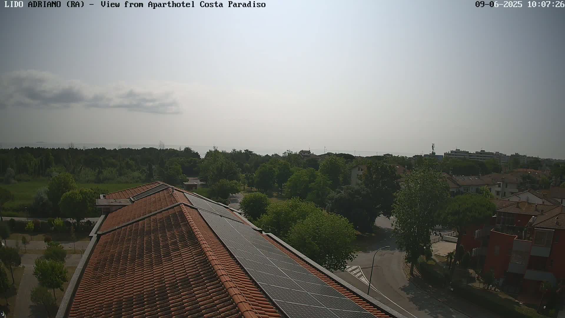A slightly hazy, sunny day overlooks a residential area with green trees, a calm sea in the distance, and a building with solar panels on its roof.