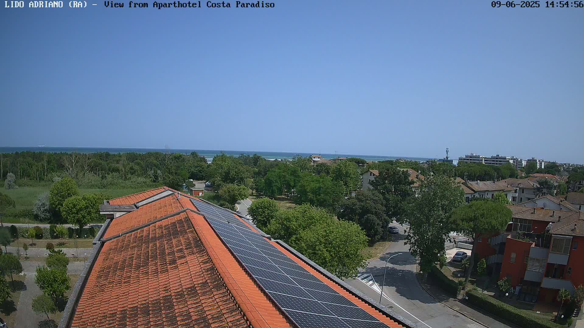 A clear, sunny day overlooking a coastal town with a building featuring solar panels on its roof, showing a calm ocean in the background.