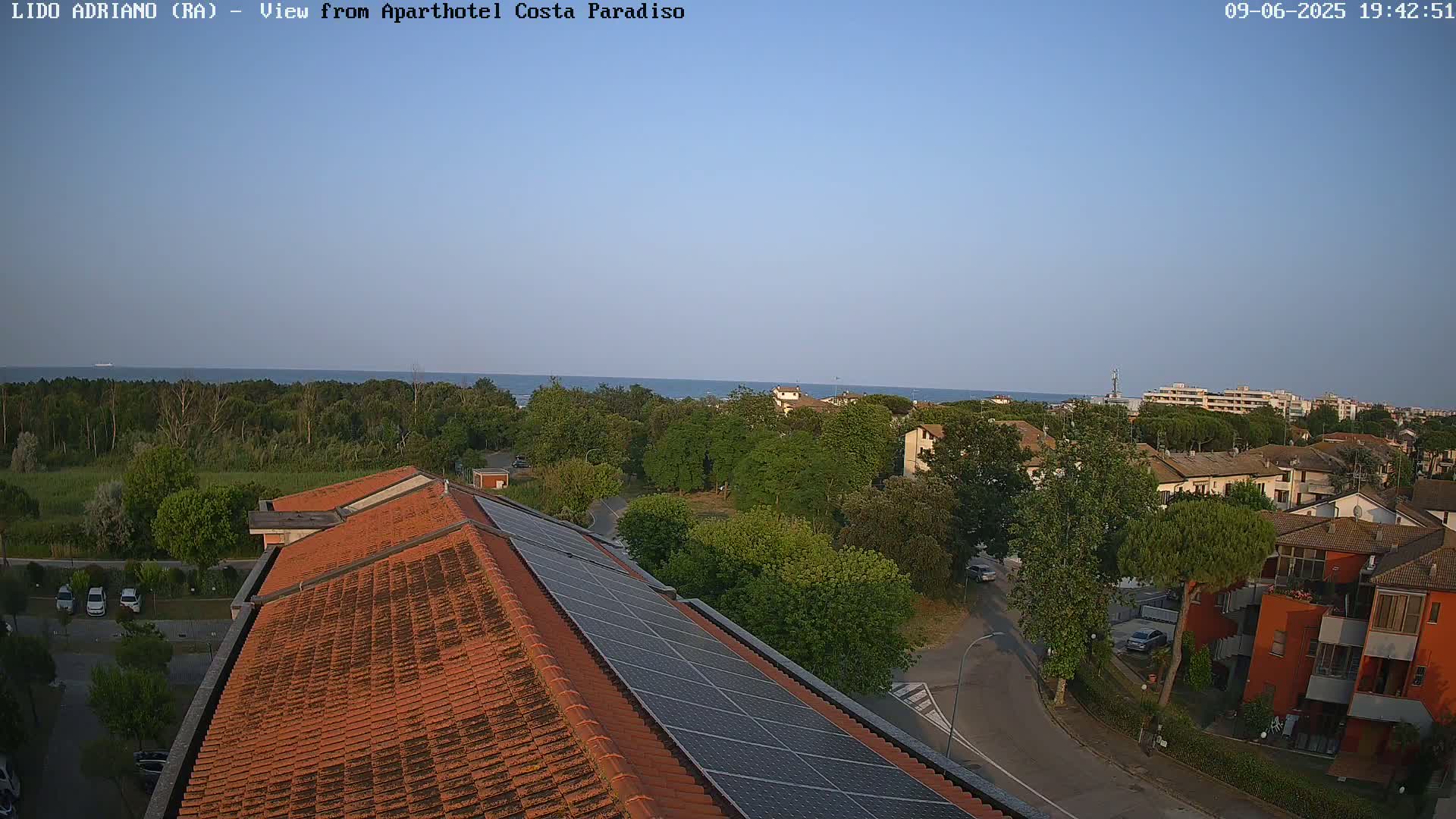 A clear, sunny view overlooking a coastal town with a red-tiled roof and solar panels, showing a calm sea in the distance.