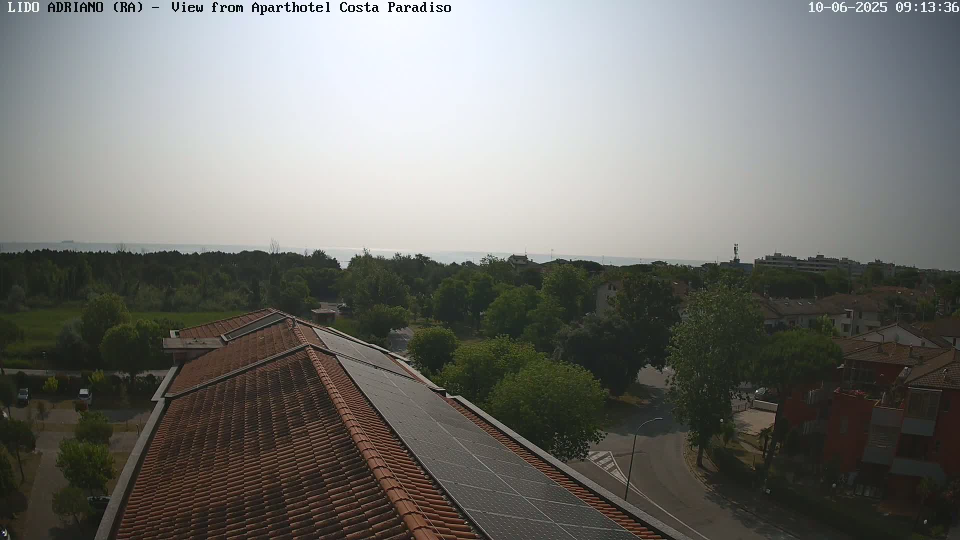 A rooftop with solar panels overlooks a residential area with green trees and a calm ocean in the distance under a hazy sky.