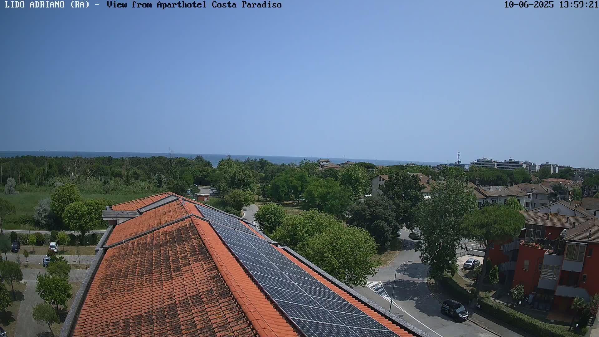 A rooftop with solar panels overlooks a residential area with green trees, a road, and a distant ocean under a clear blue sky.