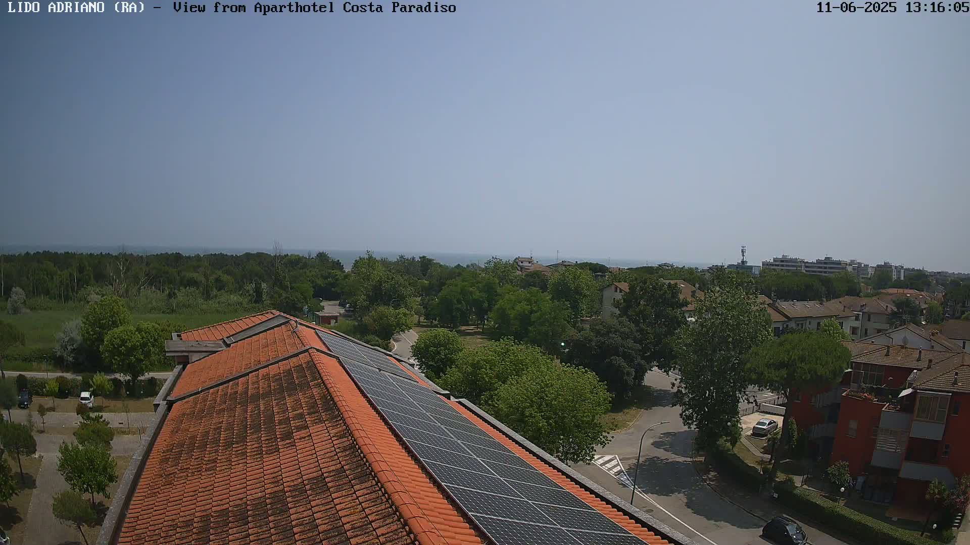 An aerial view on a sunny day shows a red-tiled roof with solar panels, overlooking a residential area with green trees and a calm sea in the distance.