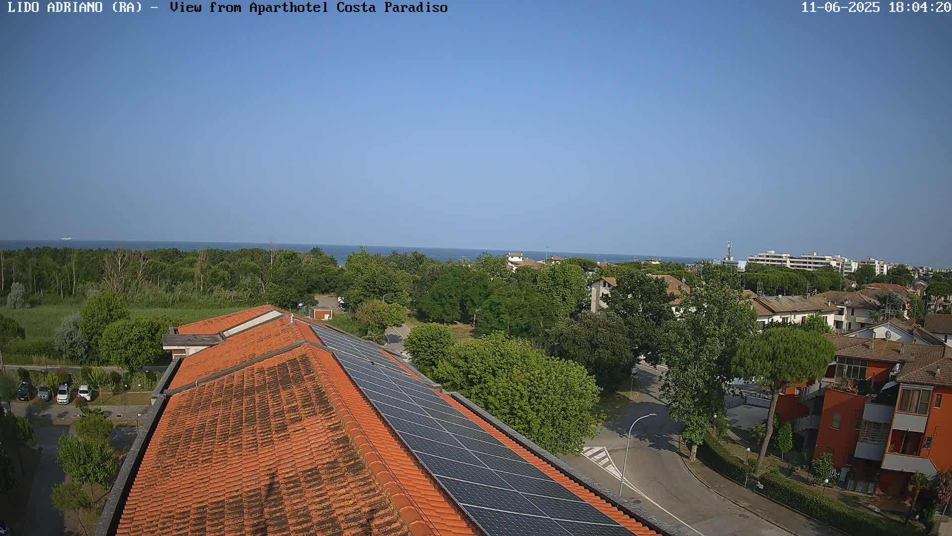 A rooftop with solar panels overlooks a residential area and a calm sea under a clear blue sky.