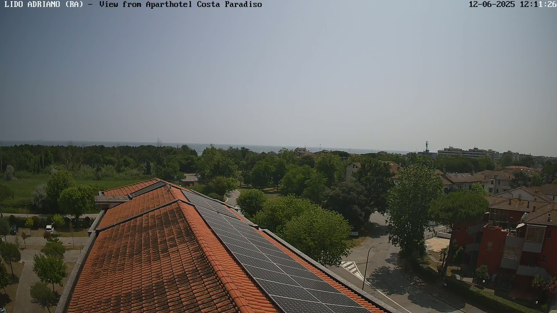 A rooftop with solar panels overlooks a residential area with green trees and a calm sea in the distance under a clear sky.