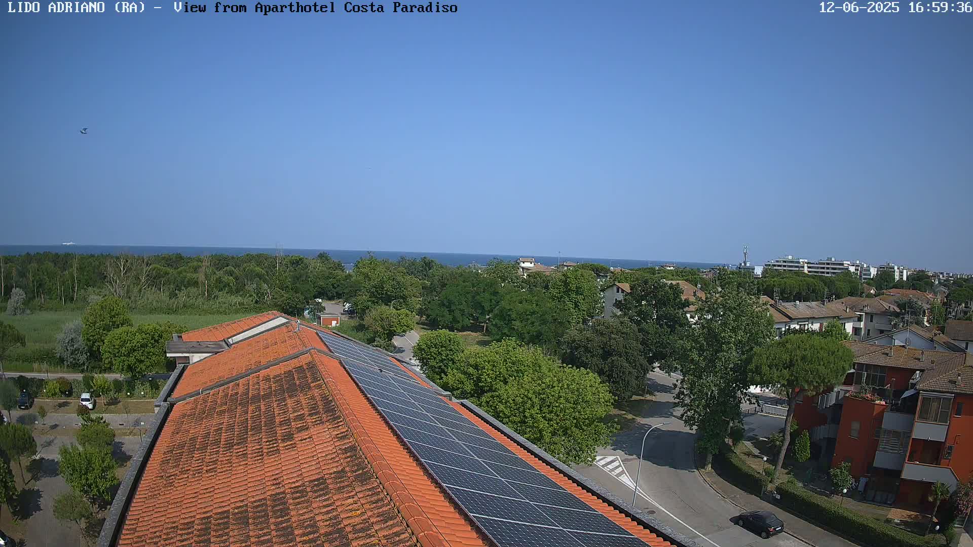 A rooftop with solar panels overlooks a residential area and a calm blue sea on a sunny day.