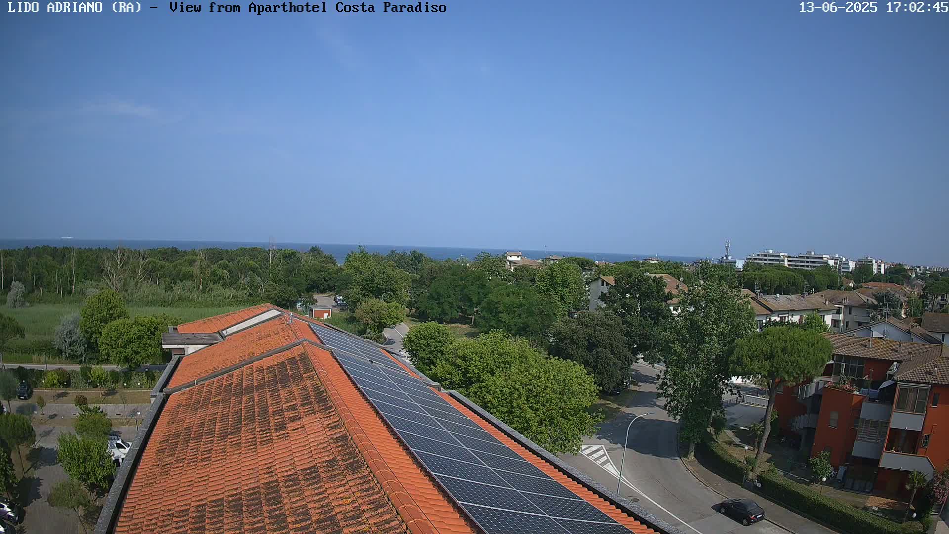 A rooftop view shows a clear blue sky over a seaside town, with a building featuring solar panels and a distant ocean visible beyond lush green trees and residential buildings.