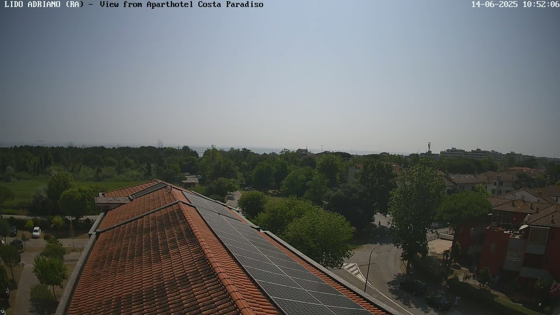 A rooftop with solar panels overlooks a residential area with green trees and a distant body of water under a clear, sunny sky.