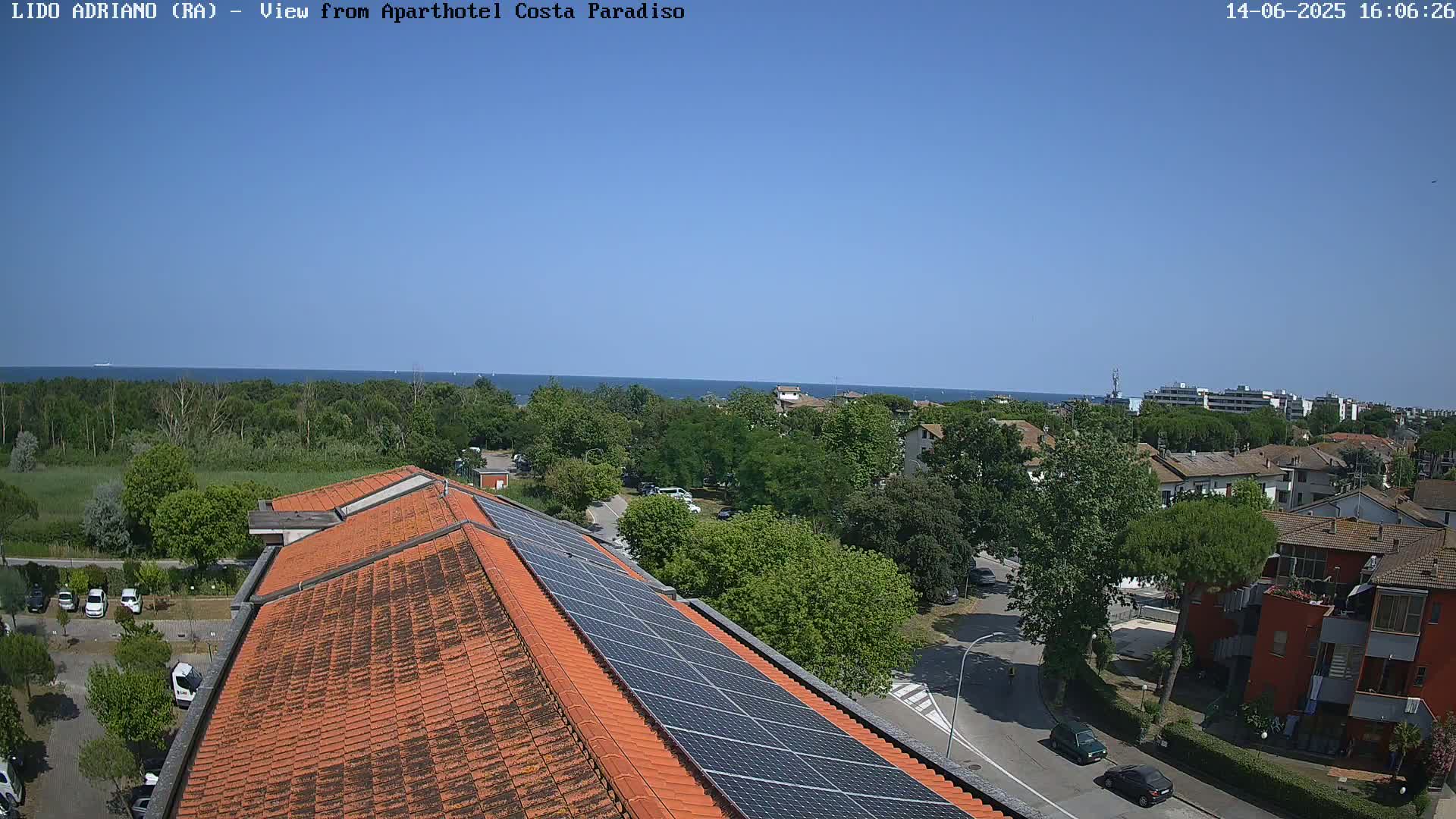 A rooftop with solar panels overlooks a coastal town on a sunny day, with a calm sea visible in the distance.