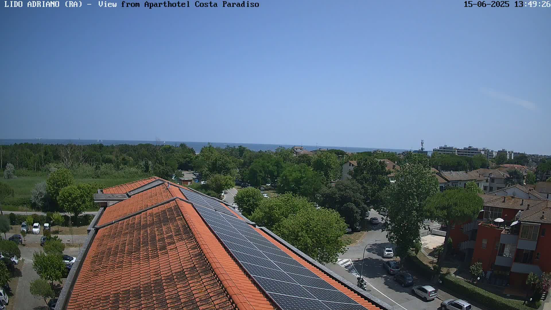 A rooftop view on a clear, sunny day shows a red-tiled roof with solar panels, overlooking a parking lot, residential buildings, and a calm ocean in the distance.