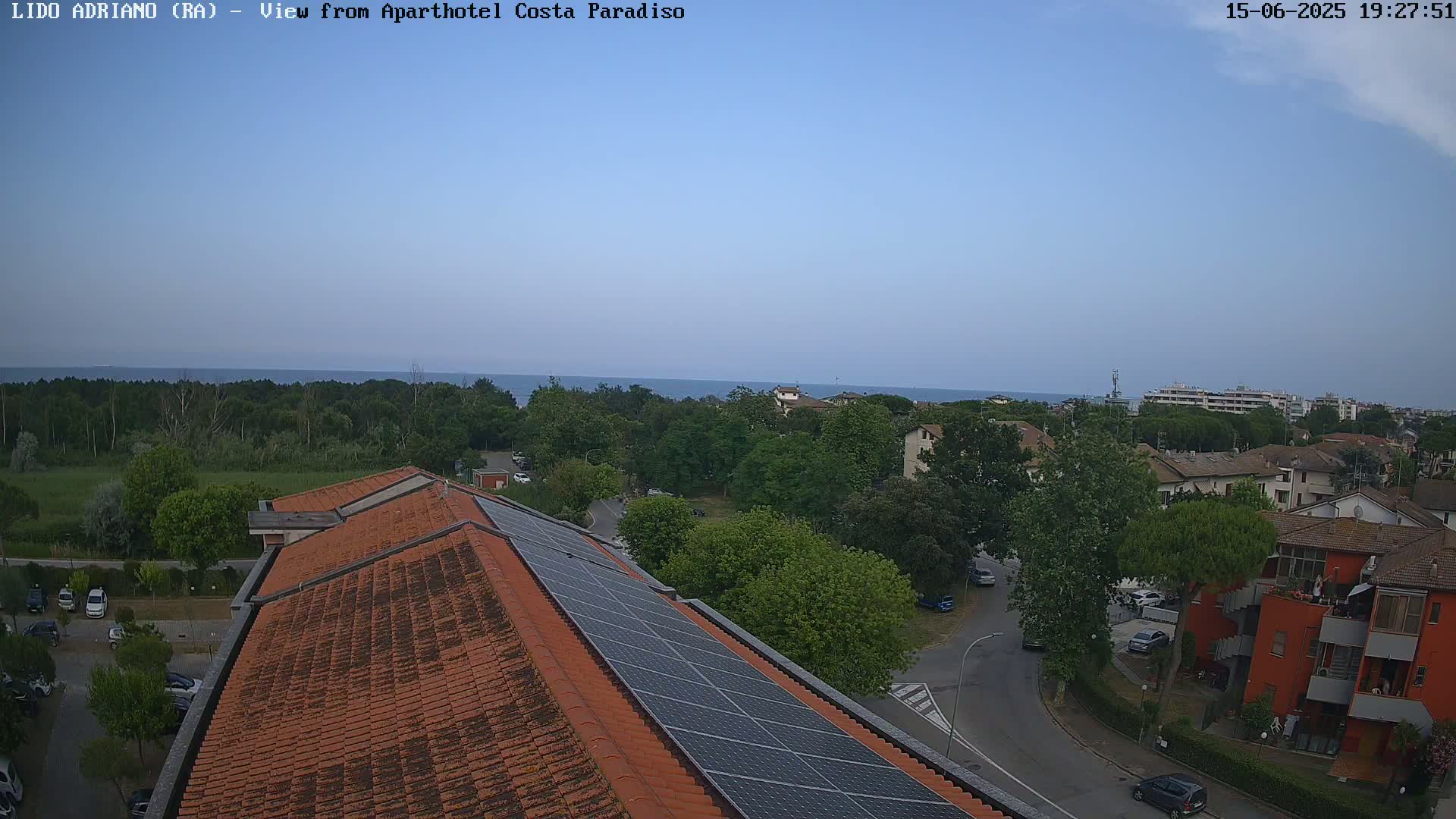 A view from a rooftop with solar panels shows a clear, calm ocean in the distance, with a residential area and green trees in the foreground.
