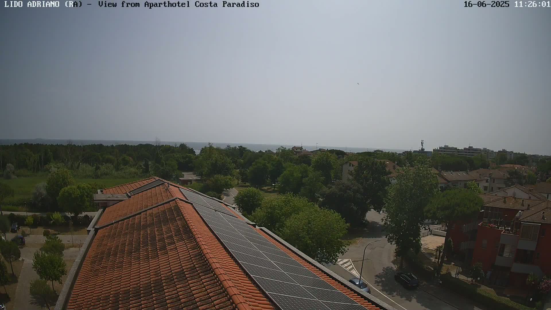 A mostly sunny day overlooking a coastal town with a red-tiled roof featuring solar panels in the foreground, and a calm sea in the distance.