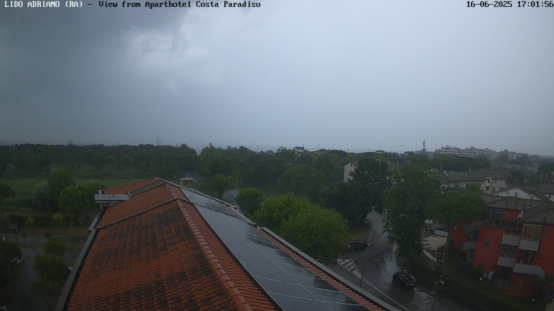 A high-angle view of a red tiled roof with solar panels during a rainy day, overlooking a residential area with lush green trees and a calm body of water in the distance under a cloudy sky.