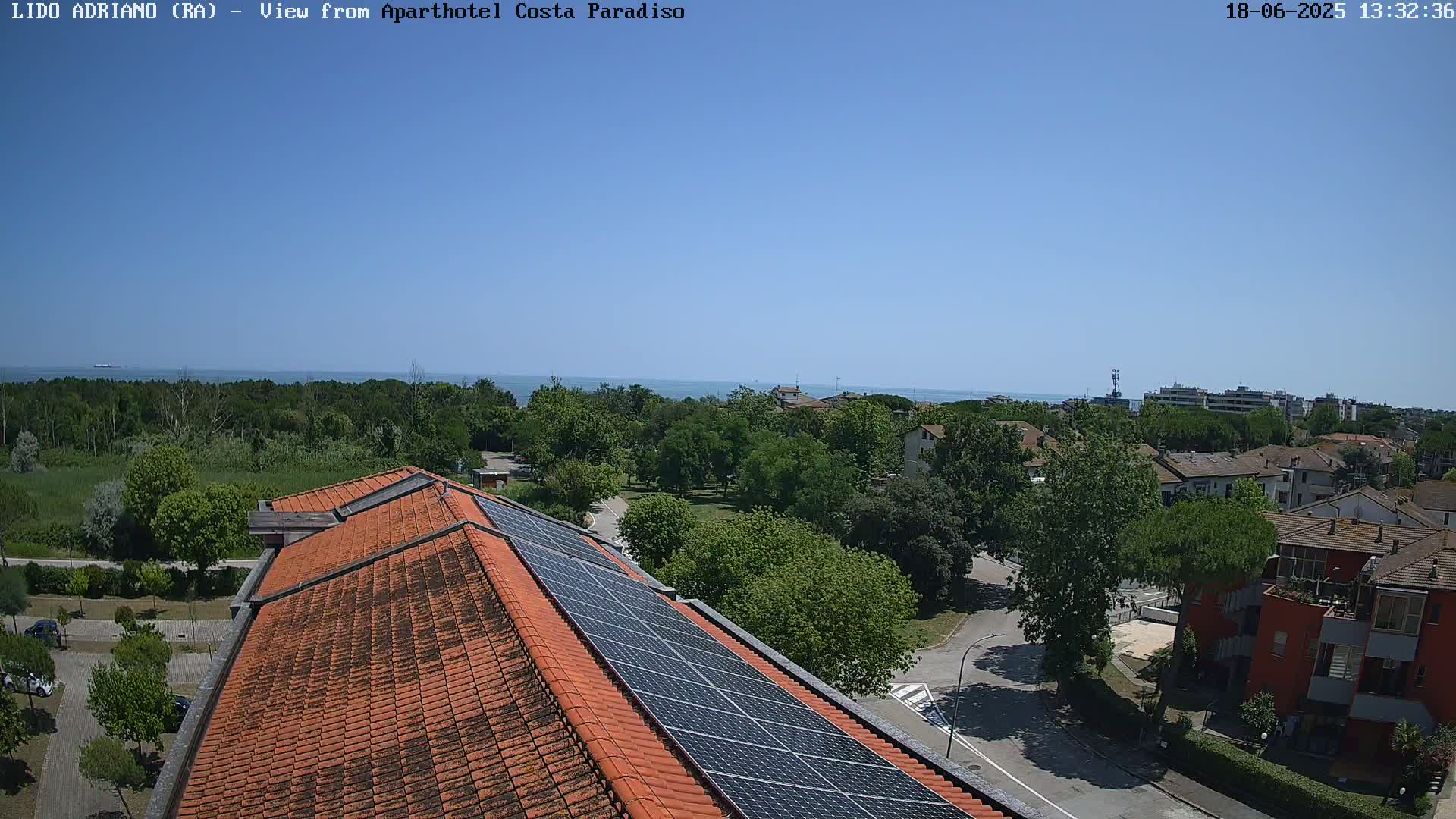 A rooftop with solar panels overlooks a residential area with green trees and a calm ocean in the distance under a clear blue sky.