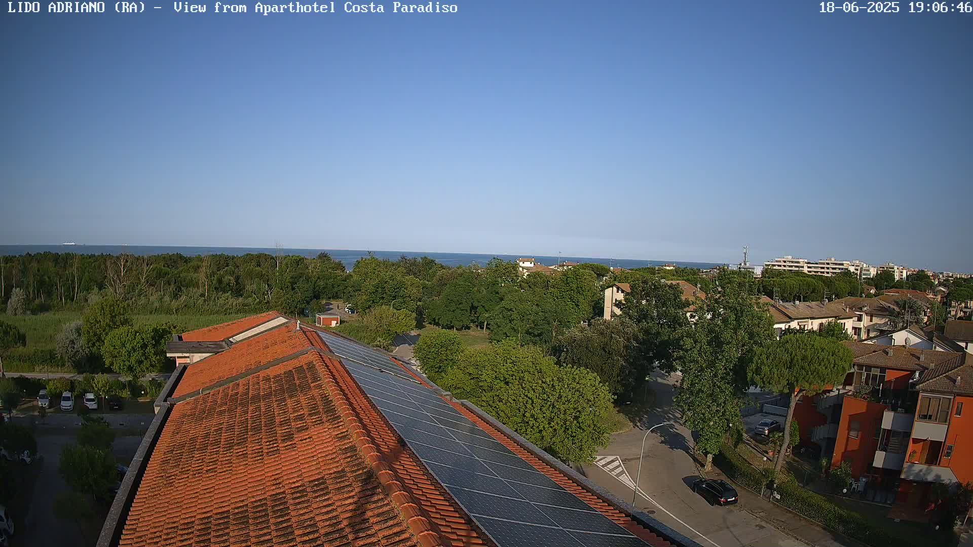 A clear, sunny day overlooking a coastal town with a red-tiled roof and solar panels in the foreground, and a calm sea in the distance.