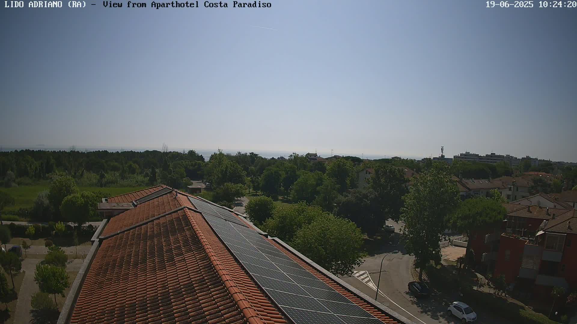 A clear, sunny day overlooks a residential area with lush green trees, a calm body of water in the distance, and a building with solar panels on its roof.